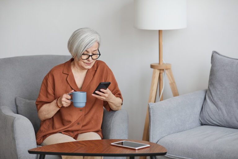 Photo of older woman drinking tea and reading smartphone while sitting on couch