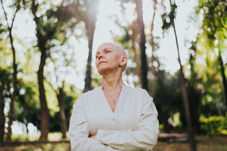 Powerful cancer patient standing in the sunshine with trees behind her.
