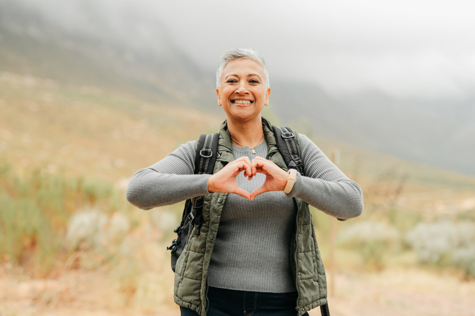 Nature hiking senior woman with love and heart sign or hand for healthy, wellness and care on adventure walk on hill, green mountain environment. Trekking person with emoji hands, portrait and smile
