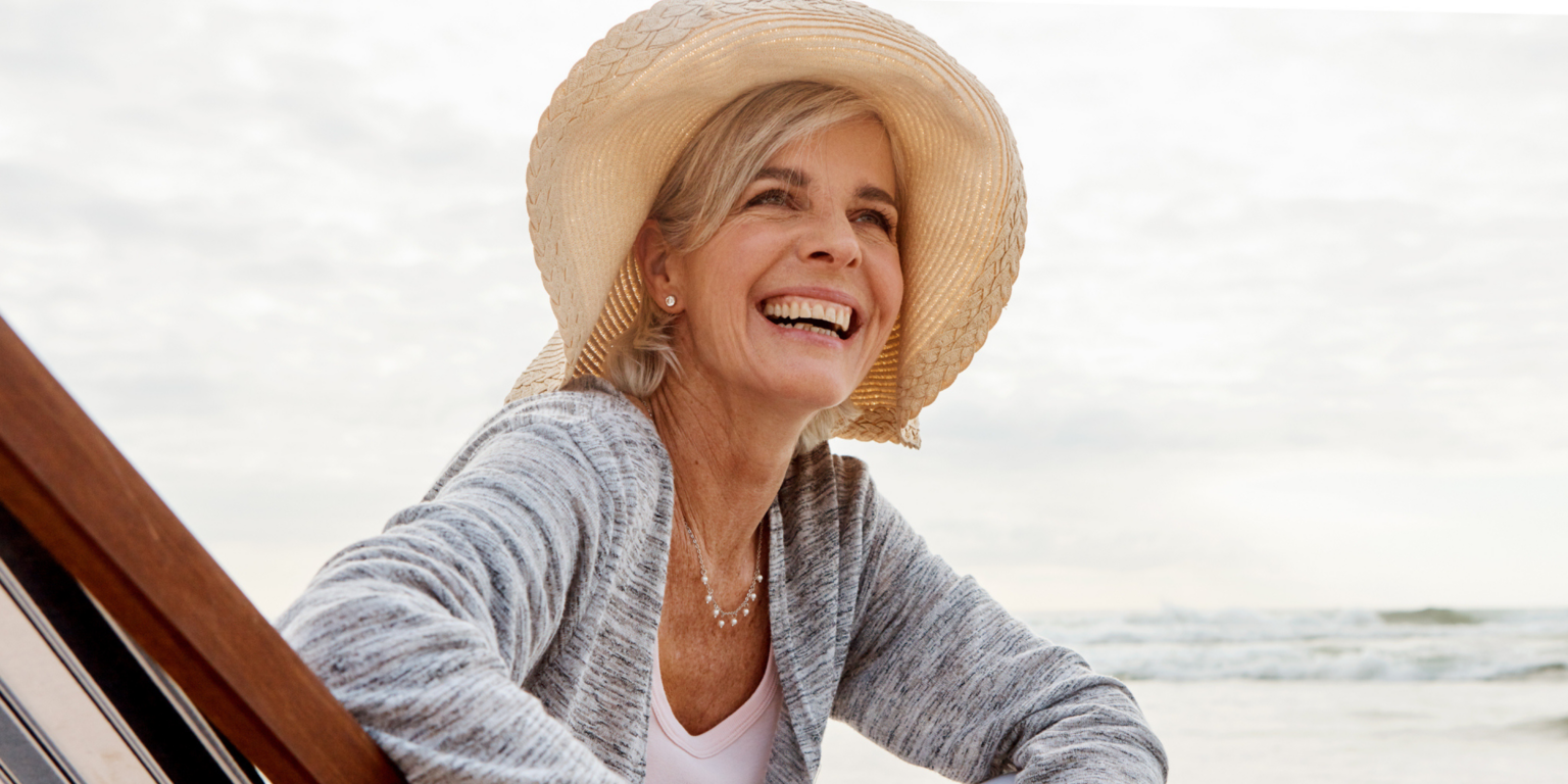 Image of a late-middle aged woman smiling, wearing a sun hat on the beach