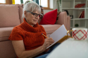 Senior woman writing in a notebook while sitting on the floor of her living room