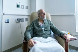 A recovering adult man cancer patient is sitting resting comfortably in a hospital cancer ward easy chair while chemotherapy IV drip medicine is administered by an array of medical equipment through a subcutaneous intravenous chemo access port temporarily embedded into his upper chest.