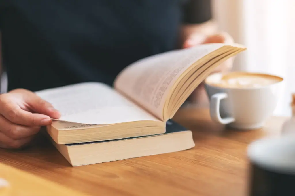 Closeup image of a woman holding and reading a vintage novel book with coffee cup on wooden table
