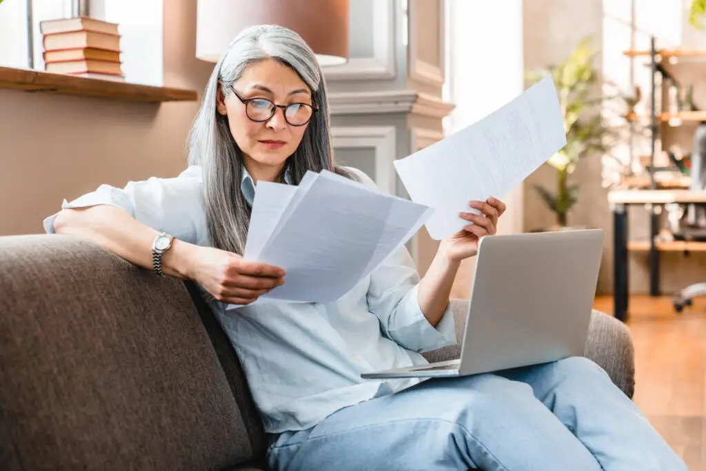 older woman sitting on couch with laptop looking at papers