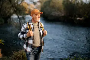 older man walking by a river with hat and backpack