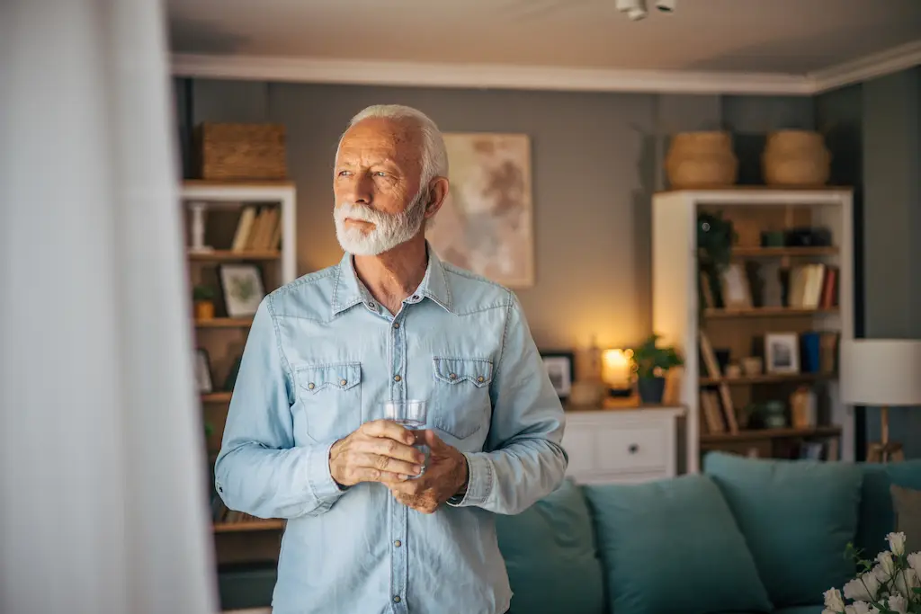 older male looking out the window with a glass of water