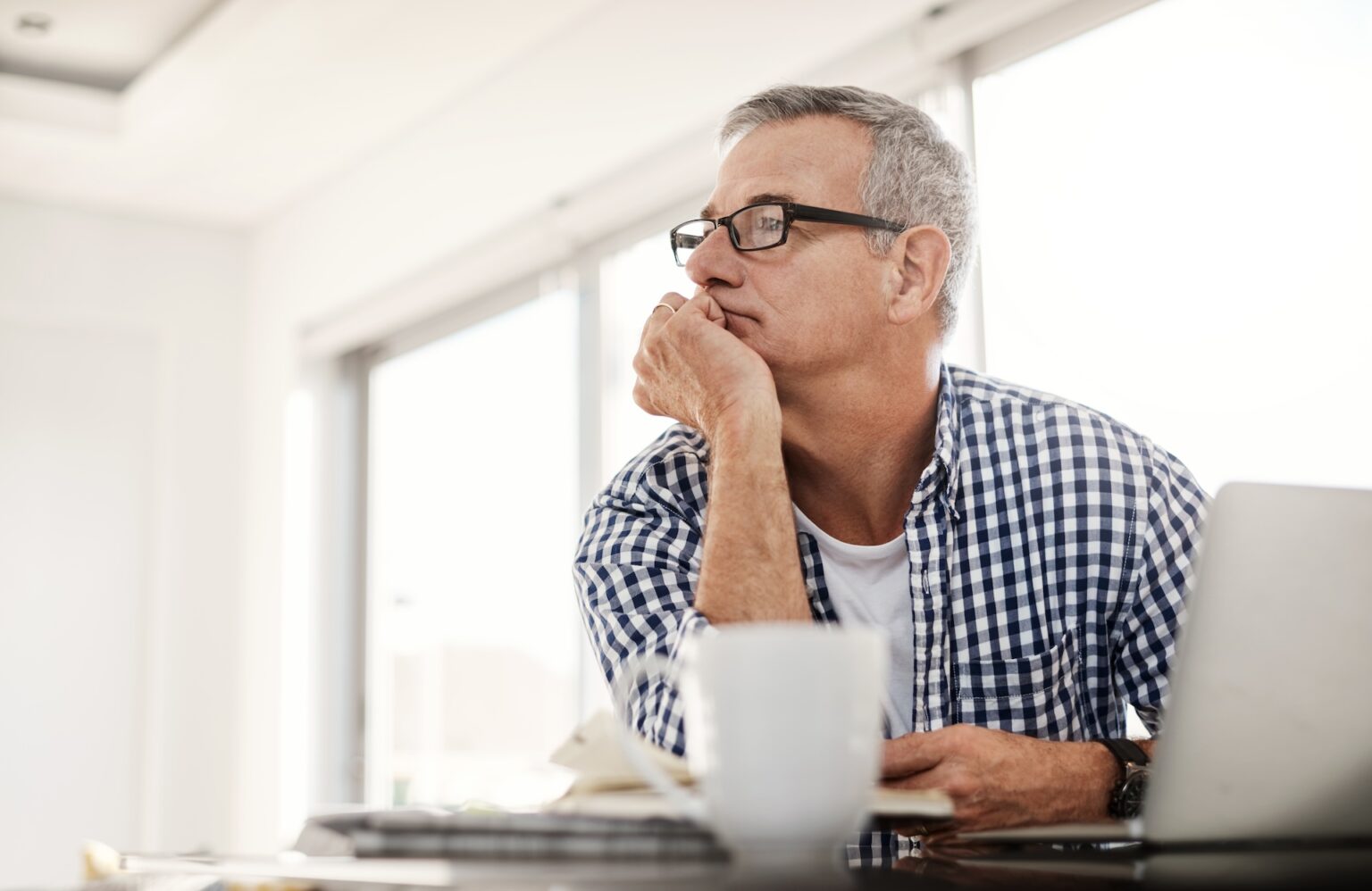 Older man sitting at table with computer and mug thinking