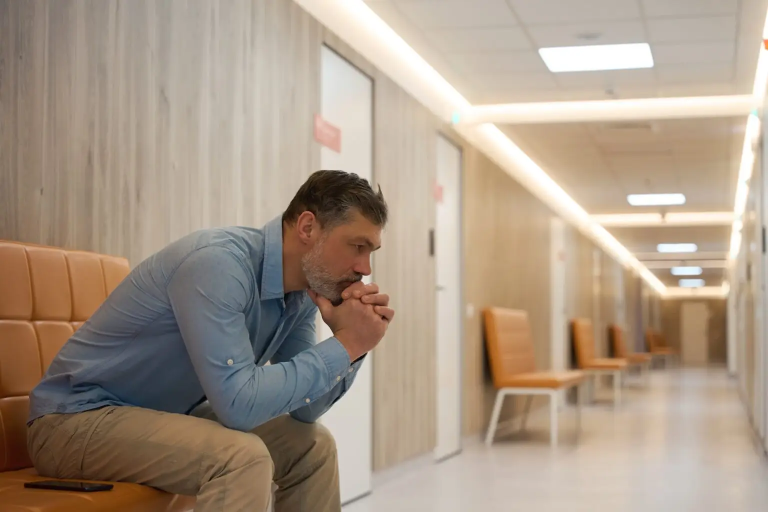 Gray-haired man with beard sits on beige sofa in lobby of medical clinic with wooden walls