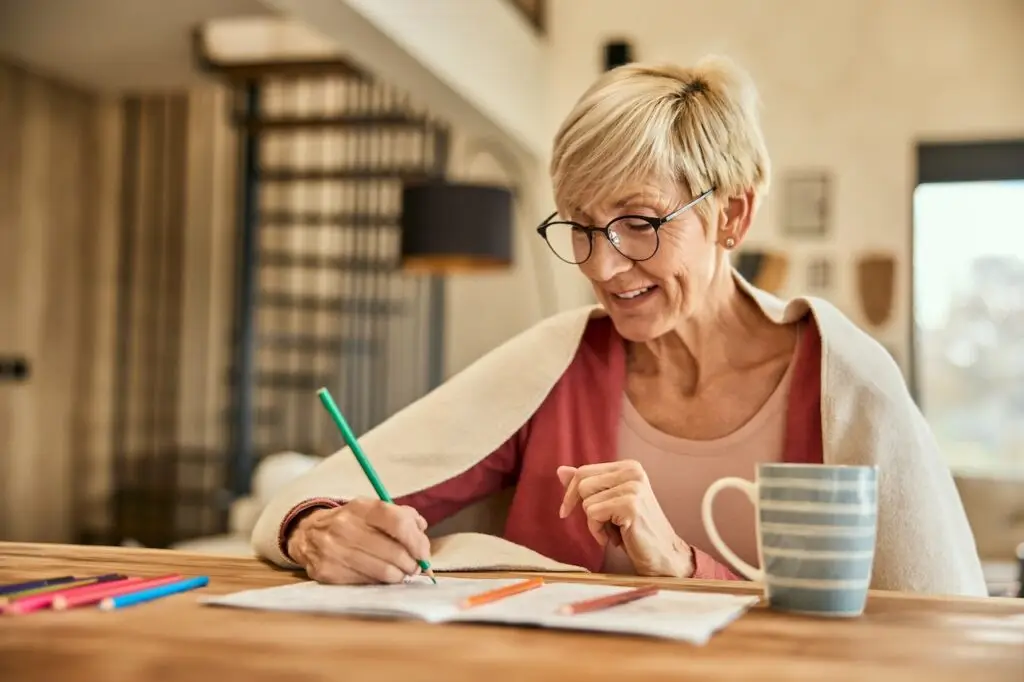 A smiling senior woman wrapped in a blanket, using a crayon and coloring the coloring book.