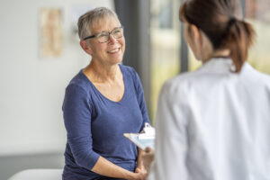 A senior woman who is dressed casually, sits up on an exam table as she attends a health check-up with her doctor.