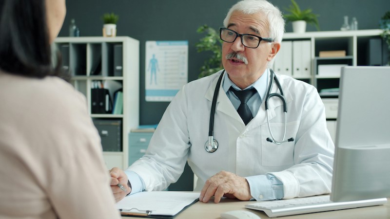 Friendly senior doctor is talking to female patient during appointment in clinic discussing disease and treatment.