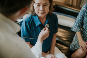 Asian Male doctor using a stethoscope to examining senior woman's lung and heartbeat during a healthcare home visit.