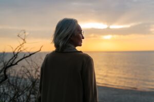 Senior woman wearing glasses and a coat standing on the seashore at sunset and looking away
