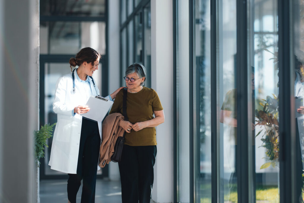 Doctor Walking With Senior Woman Patient In Hospital Corridor