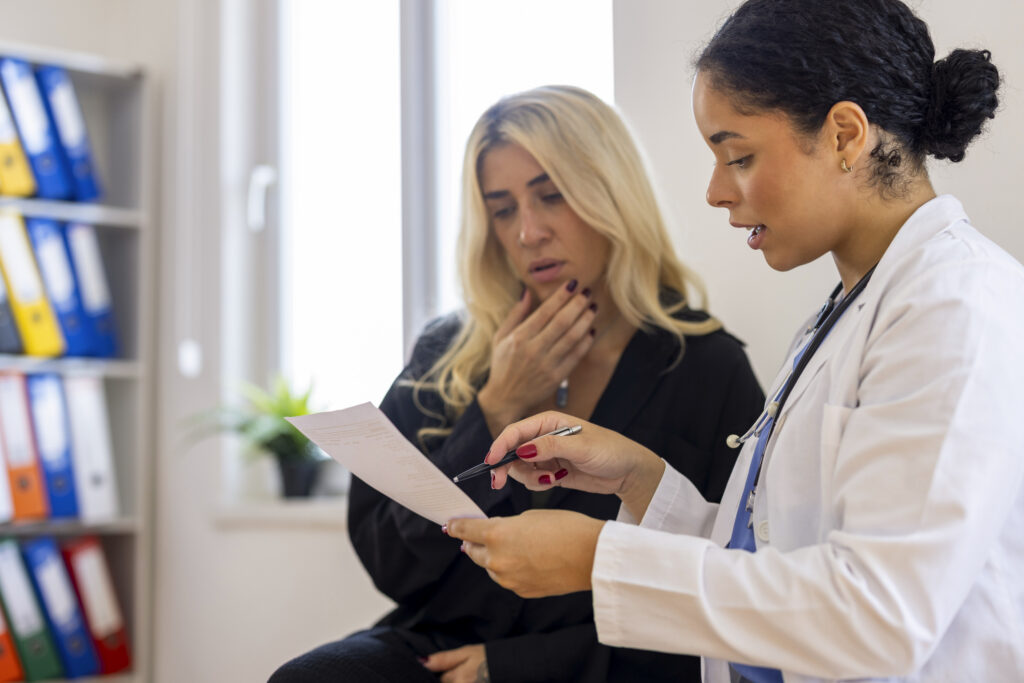 Professional female doctor with curly hair standing and showing a medical document, test result, or prescription to a blonde female patient in a clinic. The doctor is pointing at the paper with a pen, while the patient looks concerned and thoughtful, representing a serious medical consultation or diagnosis explanation.