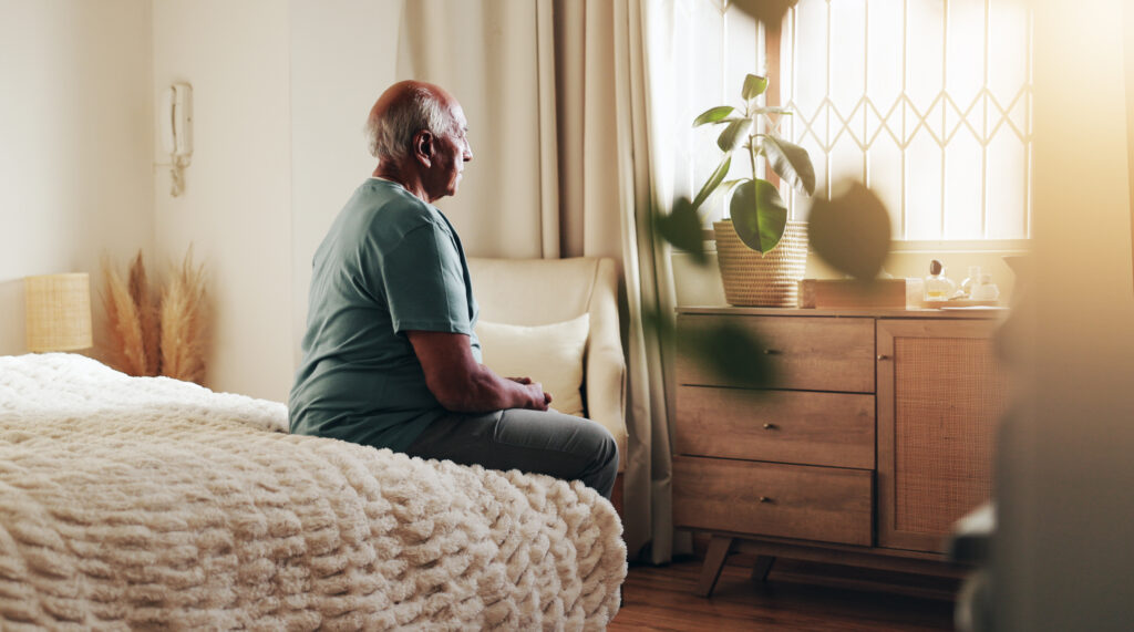 older man sitting on bed looking out the window thinking