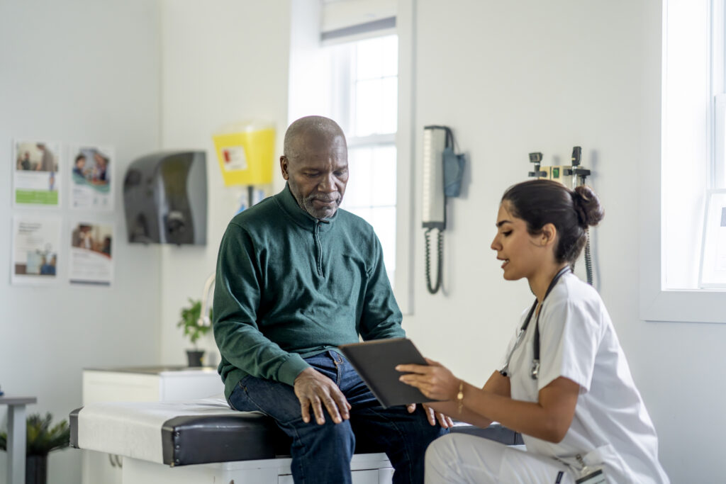 Black man sits on an examination table while a female nurse reviews information on a tablet. The scene captures a caring medical consultation in a bright clinic