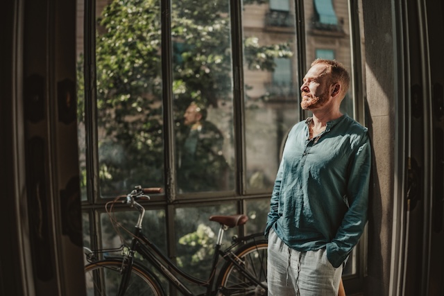 Man standing in front of a sunshine-filled window with a bicycle behind him.
